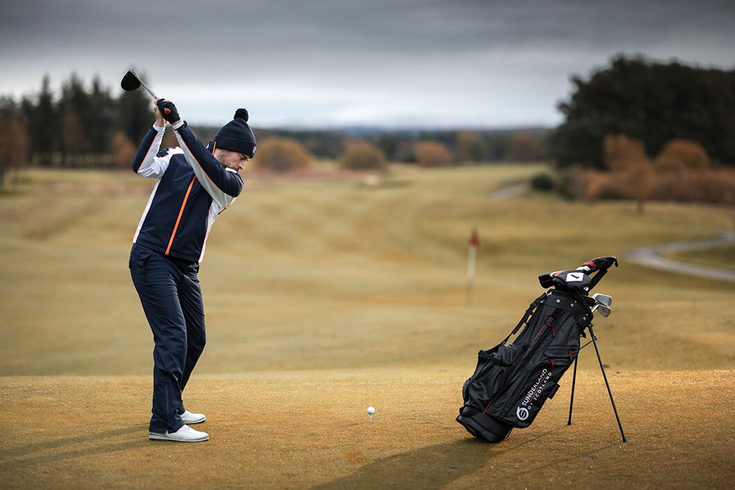 Man in navy golf waterproofs hitting drive on golf course