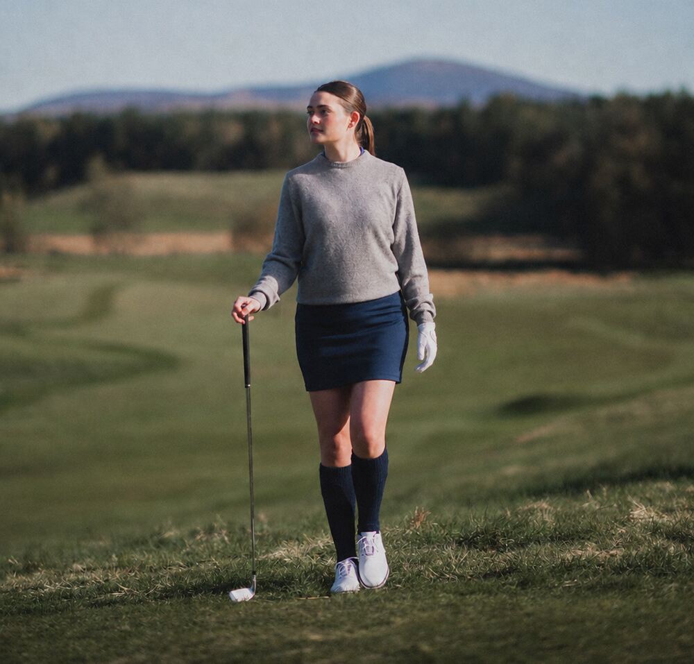 A woman on a golf course in a navy skort and grey sweater holding a club