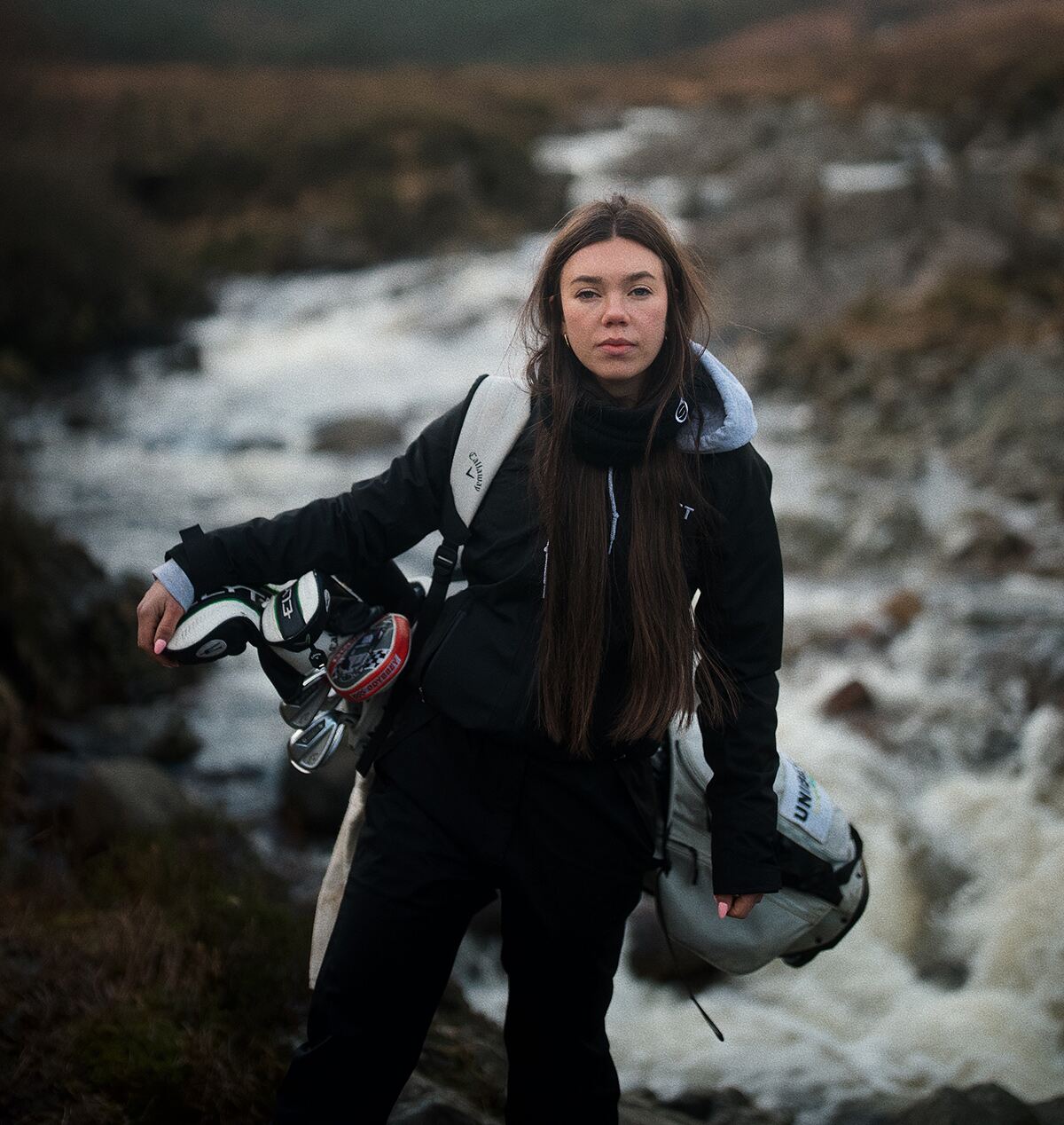 A female golfer in waterproofs carries a bag in front of a stream on a cold day winter's day
