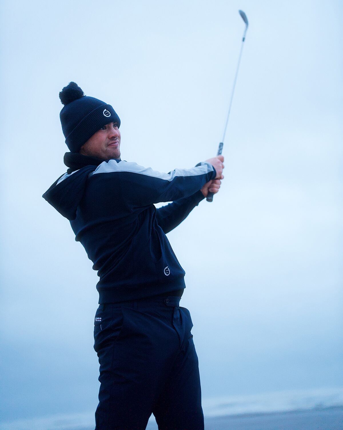 A male golfer dressed in thermal wear swings a golf club against a cold-looking grey sky